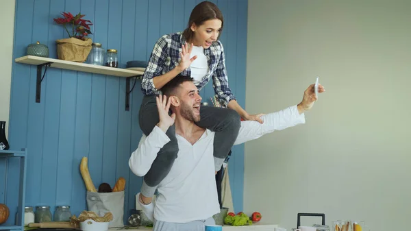 Счастливая молодая пара весело проводит время и разговаривает онлайн по видеосвязи на кухне дома. Девушка сидит на шее парня, пока он держит смартфон — стоковое фото