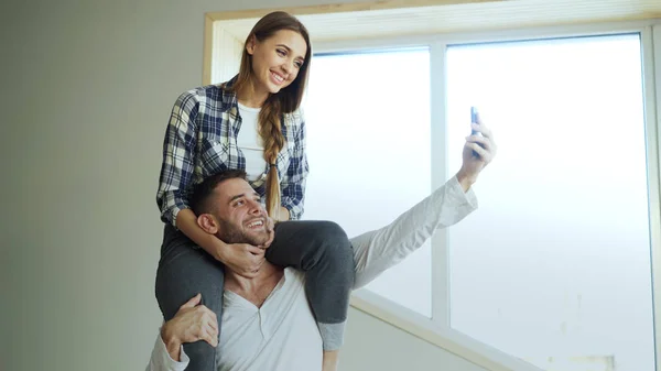 Happy νεαρό ζευγάρι έχει τη διασκέδαση και να μιλάμε σε απευθείας σύνδεση βίντεο κλήση στην κουζίνα στο σπίτι. Κοπέλα κάθεται στο λαιμό τους φίλους ενώ ο ίδιος κατέχει το smartphone — Φωτογραφία Αρχείου
