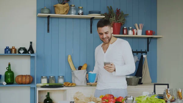 Веселый счастливый мужчина танцует и поет на кухне, а утром в социальных сетях на своем смартфоне — стоковое фото