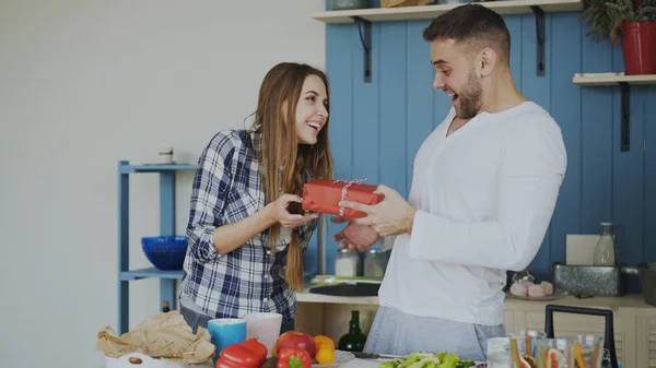 Mujer alegre sorprendiendo a su amigo boylfriend con regalo de cumpleaños en casa en la cocina mientras que él cocina el desayuno — Foto de Stock