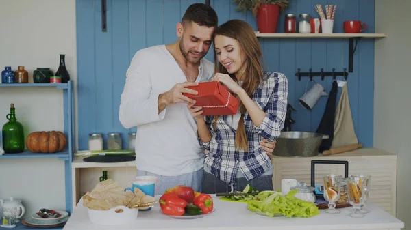 Feliz hombre alegre sorprendiendo a su novia con un regalo en casa en la cocina mientras ella cocina el desayuno — Foto de Stock