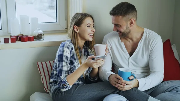 Feliz jovem casal falando e rindo enquanto sentado na cama e beber café pela manhã em casa — Fotografia de Stock