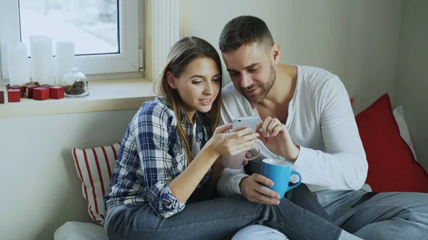 Feliz casal jovem conversando e navegando nas mídias sociais no smartphone enquanto se senta na cama e bebe café pela manhã em casa — Fotografia de Stock