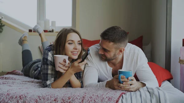 Casal sorridente assistindo TV enquanto estava deitado na cama e bebendo café em casa pela manhã — Fotografia de Stock