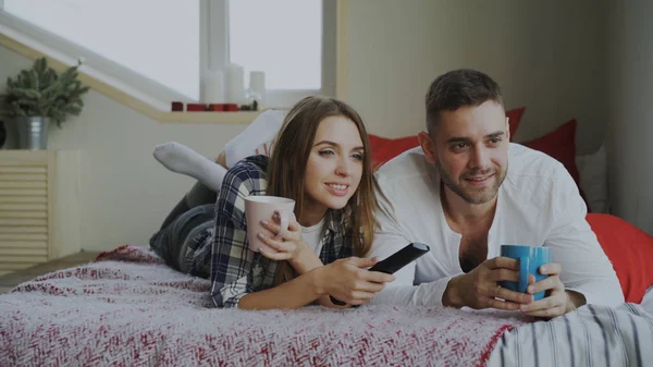 Улыбающаяся пара смотрит телевизор, лежа в постели, и пьет кофе по утрам дома — стоковое фото