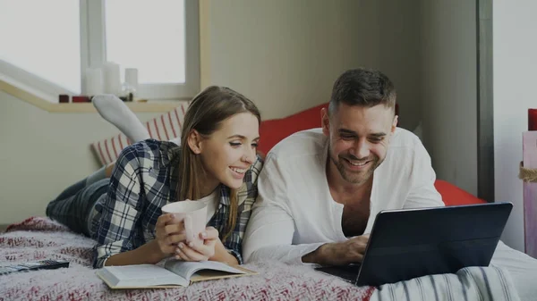 Vidám férfi és nő, beszélő és az olvasó a könyvet, és segítségével a laptop számítógép reggel ágyban fekve — Stock Fotó