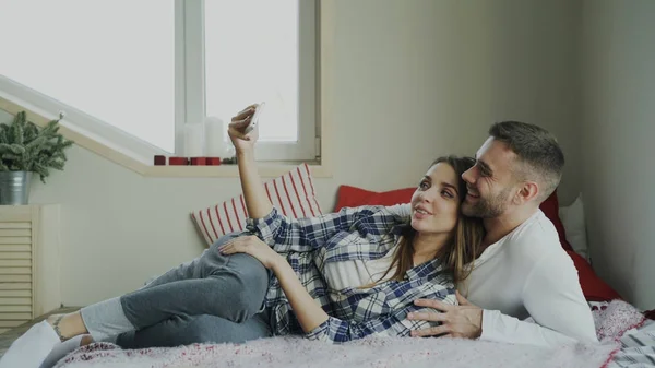 Cheerful cute and loving couple talking video call using smartphone camera and chatting with parents lying in bed at home — Stock Photo, Image