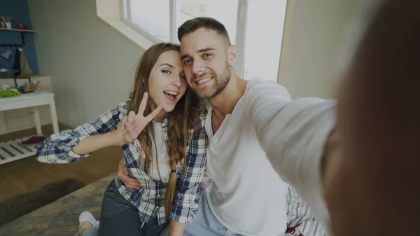 Pov junge lächelnde und schöne Paar machen Selfie-Porträt auf Smartphone-Kamera und küssen, während sie am Morgen auf dem Bett sitzen — Stockfoto