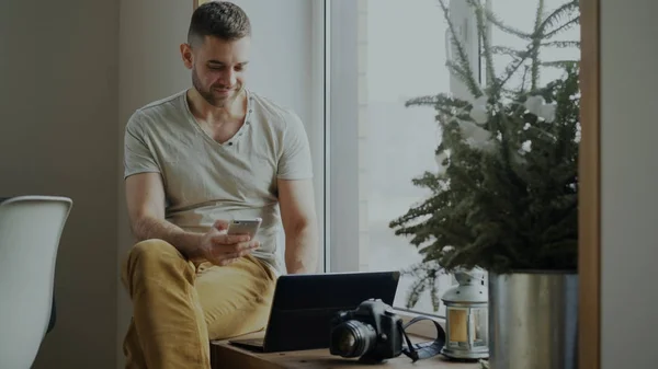 Attrayant jeune homme naviguant téléphone intelligent assis sur le rebord de la fenêtre avec ordinateur portable et caméra dans le café — Photo
