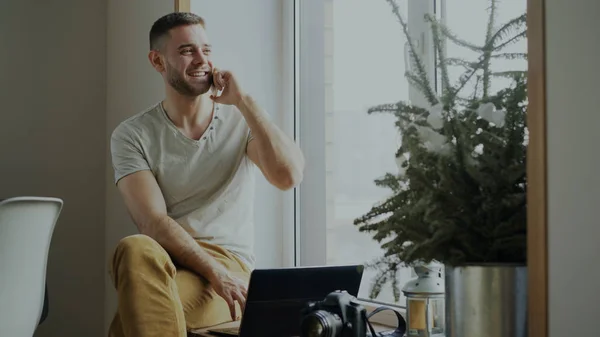 Привабливий молодий чоловік розмовляє телефоном, сидячи на підвіконні з ноутбуком і камерою вдома — стокове фото