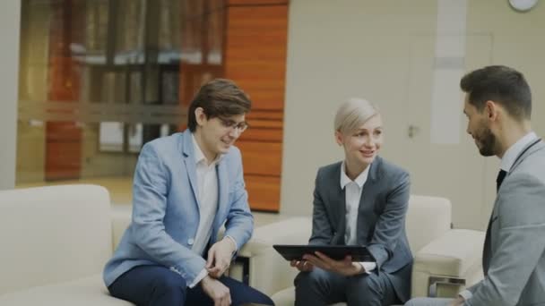 Panorámica de la mujer de negocios con tableta digital duscussing con colegas de negocios masculinos sentados en el sofá en la oficina moderna — Vídeo de stock