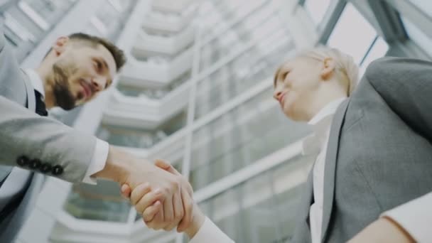 Disparo de bajo ángulo de hombre de negocios barbudo estrechando la mano y hablando con una colega de negocios en traje en el vestíbulo del edificio de oficinas moderno — Vídeo de stock