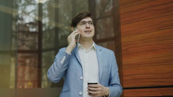 Panorámica de un joven empresario hablando teléfono inteligente con taza de café en la oficina moderna — Vídeos de Stock