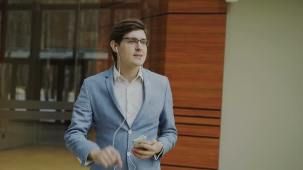 Feliz hombre de negocios escuchando música con auriculares bailando y caminando en el vestíbulo de la oficina moderna — Vídeos de Stock