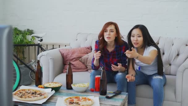 Dos amigas multiétnicas viendo partidos deportivos en la televisión juntas en casa mientras beben cerveza y comen bocadillos — Vídeos de Stock