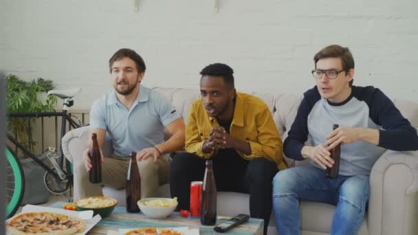 Група молодих друзів-чоловіків, які дивляться спортивний матч по телевізору разом, п'ють пиво і їдять закуски вдома — стокове відео