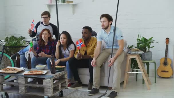 Multi grupo étnico de amigos fãs de esportes com bandeiras nacionais norueguesas assistindo campeonato de hóquei na TV juntos animando sua equipe favorita em casa — Vídeo de Stock