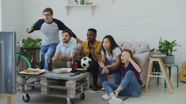 Grupa młodych przyjaciół oglądania meczu piłki nożnej w telewizji razem jedzenia przekąski i picia piwa w domu. Ludzie są zadowoleni z ich zwycięstwo gospodarzy, ale dziewczyny rozczarowany — Wideo stockowe