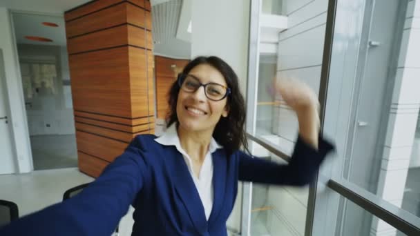 Фотография молодой бизнесвумен в костюме, делающей селфи, держа в руках смартфон и веселясь в современном офисе — стоковое видео