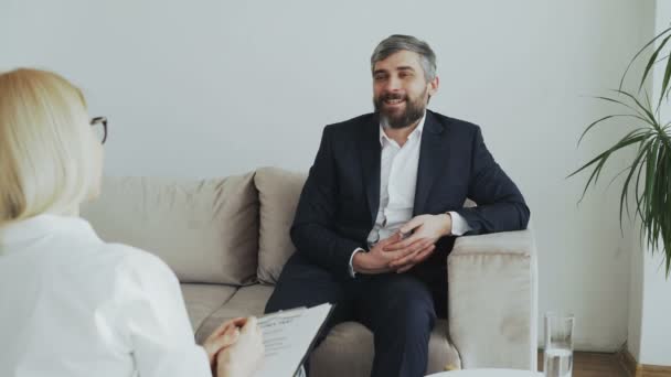 Улыбающийся бизнесмен сидит на диване и разговаривает с женщиной-психологом в офисе — стоковое видео