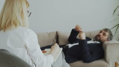 Psikoterapist veya psikolog ofisinde kanepede yatan işadamı ile konuşurken not yazma