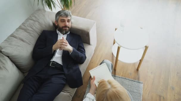 プロの女性の心理や彼女のオフィスで心理学者と彼の問題について話しているソファの上に横になっている男のトップ ビュー — ストック動画