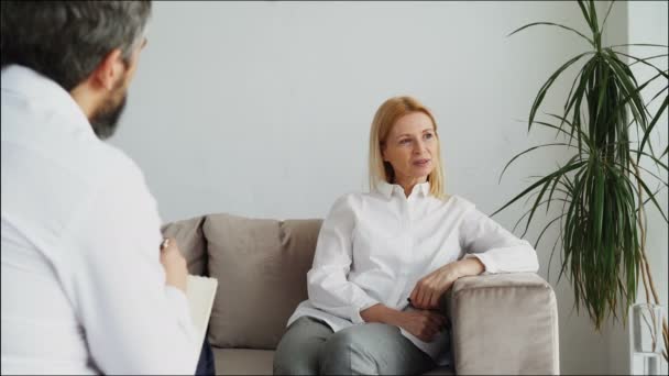 Мужчина-психоаналитик слушает взрослую пациентку и пишет заметки в офисе психолога — стоковое видео