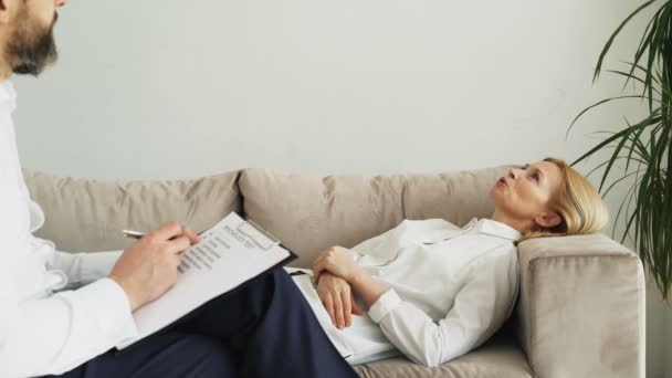 Erwachsene Frau, die auf der Couch liegt und über ihre Probleme mit einem professionellen männlichen Psychotherapeuten oder Psychologen in seinem Büro spricht — Stockvideo