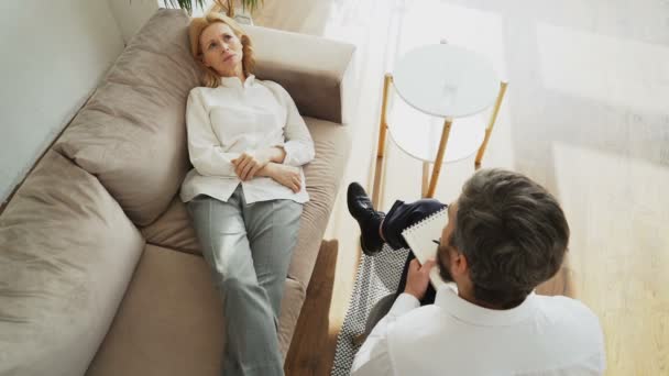 Вид зверху засмучена жінка лежить на дивані, говорячи про свої проблеми з професійним чоловіком психотерапевтом або психологом в його офісі — стокове відео
