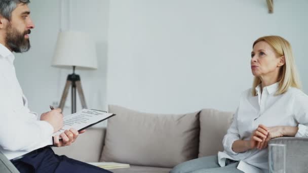 Male experienced psychologist talking and listening to upset female patient and writing notes on clipboard in his office — Stock Video