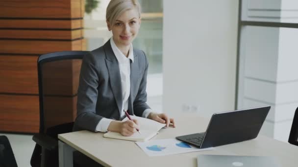 Portrait of Attractive blonde businesswoman sitting at table writing in notebook smiling into camera in modern office — Stock Video