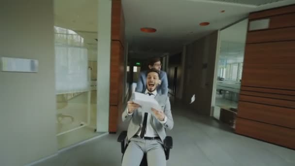 Dolly Shot von zwei verrückten Geschäftsleuten, die Bürostuhl fahren und Papiere hochwerfen, während sie Spaß in der Lobby des modernen Business Centers haben — Stockvideo