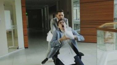 İki deli İşadamlarından ofis koltuğu sürme ve modern iş merkezi lobisinde eğlenirken kağıtları kusuyor ağır çekim dolly vurdu