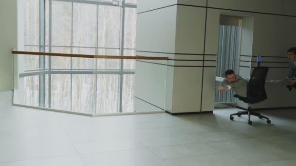 Повільна рухова панель знімала двох смішних бізнесменів, які їздять на офісному стільці, розважаючись у вестибюлі сучасного бізнес-центру — стокове відео
