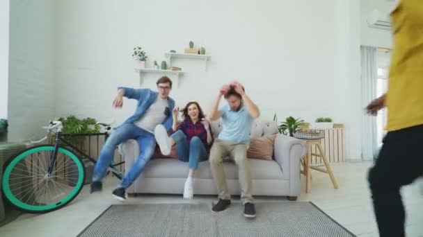Jóvenes amigos tienen corriendo y saltando en el sofá y luego sonriendo y mostrando los pulgares hacia arriba todos juntos en casa — Vídeos de Stock