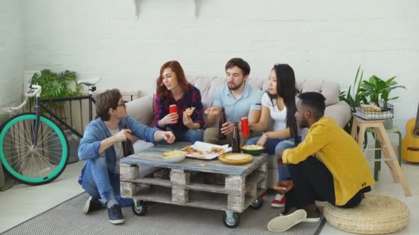 Счастливые друзья едят пиццу и празднуют вечеринку звон бутылок с пивом и газировкой сидя дома на диване — стоковое видео