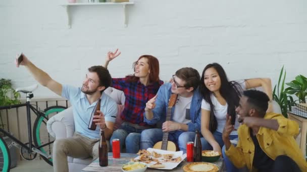 Felices amigos estudiantes tomando selfie en el teléfono inteligente y posando mientras hacen fiesta con cerveza y pizza en un alojamiento compartido en el interior — Vídeo de stock