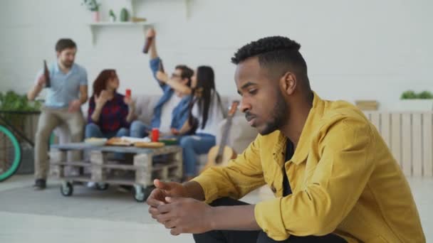 Nuori afrikkalainen amerikkalainen mies tuntee järkyttynyt ja eristäytynyt, kun hänen ystävänsä juhlivat puolue kotona sisätiloissa — kuvapankkivideo
