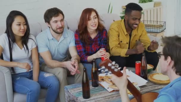 Felices jóvenes amigos tienen fiesta en el apartamento compartido y cantan juntos mientras su amigo toca la guitarra en casa — Vídeo de stock