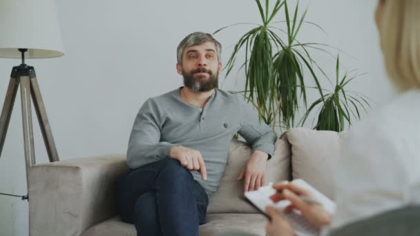 Sorrindo homem adulto sentado no sofá e falando com psicanalista do sexo feminino em seu escritório dentro de casa — Vídeo de Stock