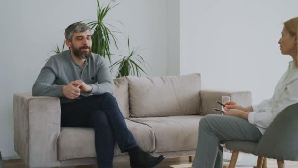 Взрослый бородатый мужчина говорит о своих проблемах психологу в ее кабинете в помещении — стоковое видео