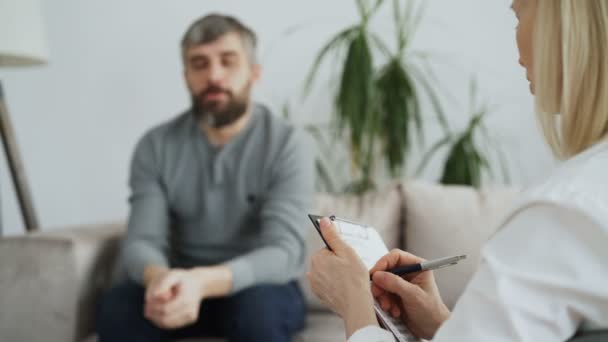 Крупный план психотерапевта или психолога, пишущего заметки во время разговора со взрослым мужчиной, сидящим на диване в ее офисе — стоковое видео