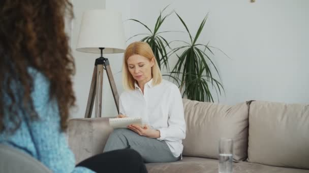 女性经验丰富的心理学家谈论和听不安的妇女病人和写笔记在剪贴板在她的办公室室内 — 图库视频影像