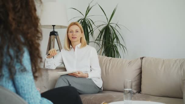 Femme psychologue expérimenté parler et écouter bouleversé fille patiente et d'écrire des notes sur presse-papiers dans son bureau à l'intérieur — Video