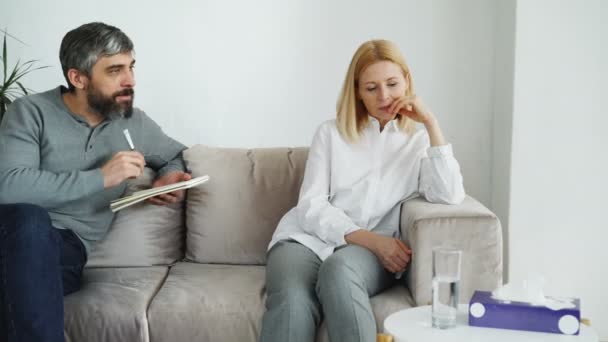 Psicólogo experimentado masculino hablando y escuchando a paciente mujer deprimida y escribiendo notas en su oficina — Vídeos de Stock