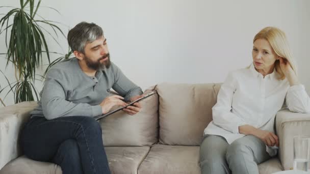 Чоловік досвідчений психолог розмовляє і слухає засмучену пацієнтку і пише нотатки на буфері у своєму офісі — стокове відео