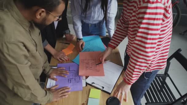 Tilt up of Group of multiethnic young coworkers watching papers with building plan standing around table in office — Stock Video