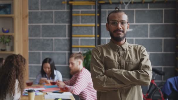 Портрет молодого африканского бизнесмена в рубашке и очках, улыбающегося и смотрящего в камеру, в то время как его коллеги работают в современном офисе в помещении — стоковое видео