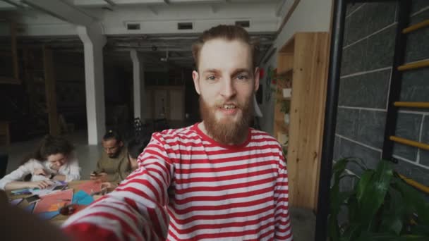 Primer plano del hombre hipster sosteniendo el teléfono inteligente y tienen videollamada en vivo mientras sus colegas trabajan en la oficina moderna — Vídeos de Stock