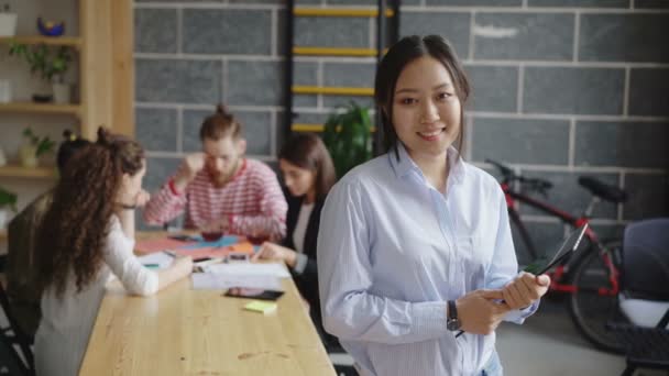 Портрет молодої азіатки-підприємниці, що тримає цифровий планшет, дивиться на камеру і посміхається на зайнятому стартовому офісі — стокове відео